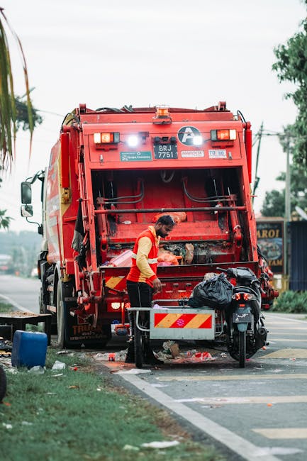 A worker wearing a yellow and red high-visibility vest is standing next to the rear of a large red waste collection truck, which is parked on the side of a street. The truck has its rear loading hatch