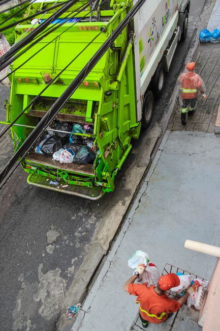 A worker wearing a yellow and red high-visibility vest is standing next to the rear of a large red waste collection truck, which is parked on the side of a street. The truck has its rear loading hatch