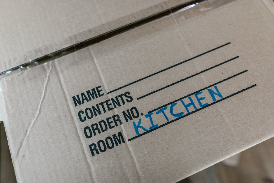 A young woman sitting on a wooden floor inside a home surrounded by numerous unpacked cardboard boxes of various sizes, some sealed with red packing tape and others open. She is dressed casually in a