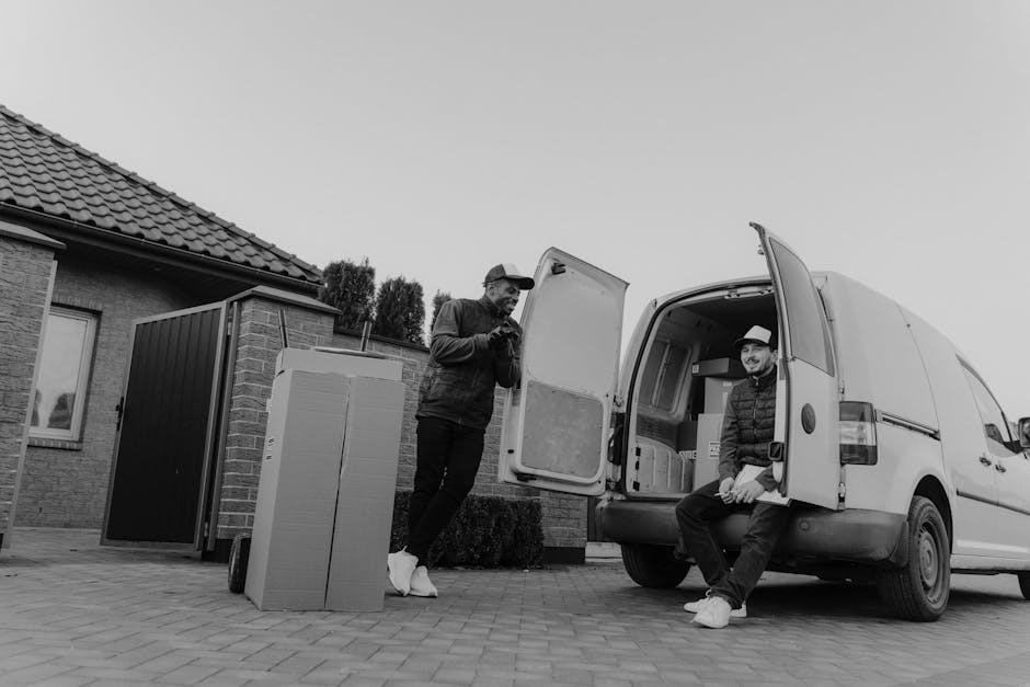 Two men are engaged in a home relocation task outside a residential property, preparing a white cargo van for furniture transport and packing. One man, standing beside the open rear door of the van, is holding a small object and appears to be inspecting or organizing items. The other man, seated on the edge of the open van door, is smiling and wearing a hat, with his hands resting on his knees. In front of the van, several cardboard boxes and a large black plastic crate are placed on the paved driveway, ready for loading. The van is positioned near the front of a house with a tiled roof and brick walls, with a nearby garden and hedge visible in the background. The scene is set during daytime with natural lighting, capturing an organized process of furniture transport, packing, and moving logistics, typical of a professional removals service such as Man and Van Harringay.