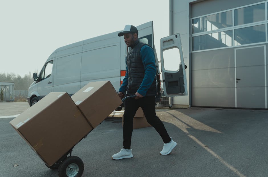 A man wearing a dark jacket, cap, and white sneakers is engaged in home relocation activities outside a modern building with large glass windows. He is lifting a cardboard box with handles onto a two-wheeled hand truck, which already holds another similar box. The boxes are sealed with packing tape and appear to contain household items for moving. Behind him, a white delivery van with its sliding door open is parked, ready for furniture transport, with the loading area adjacent to the building’s entrance. The scene suggests a loading process as part of a house removals operation involving packing, handling, and vehicle loading. The environment is well-lit with natural daylight, emphasizing the logistical aspect of packing and moving services that Man and Van Harringay offers as part of their professional removals service category.
