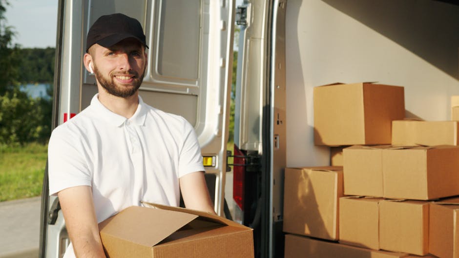 Two men are engaged in a home relocation task outside a residential property, preparing a white cargo van for furniture transport and packing. One man, standing beside the open rear door of the van, is holding a small object and appears to be inspecting or organizing items. The other man, seated on the edge of the open van door, is smiling and wearing a hat, with his hands resting on his knees. In front of the van, several cardboard boxes and a large black plastic crate are placed on the paved driveway, ready for loading. The van is positioned near the front of a house with a tiled roof and brick walls, with a nearby garden and hedge visible in the background. The scene is set during daytime with natural lighting, capturing an organized process of furniture transport, packing, and moving logistics, typical of a professional removals service such as Man and Van Harringay.