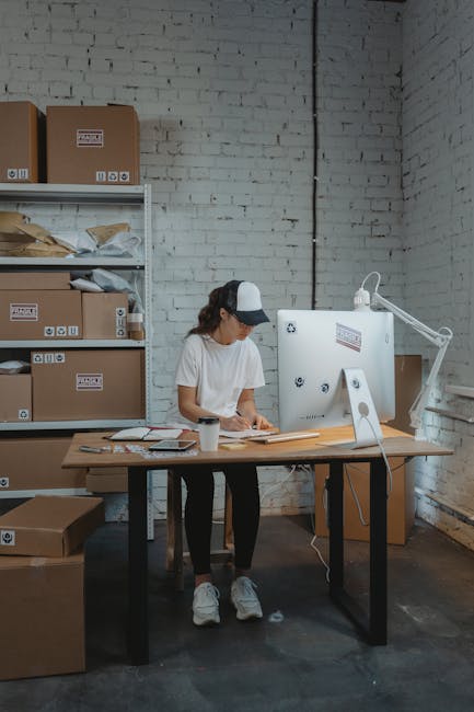 A woman wearing a white t-shirt, dark pants, and a baseball cap is standing at a wooden desk inside a warehouse or storage room with white brick walls. She is engaged in packing or organizing items, u