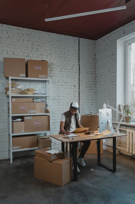 A woman wearing a white t-shirt, dark pants, and a baseball cap is standing at a wooden desk inside a warehouse or storage room with white brick walls. She is engaged in packing or organizing items, u